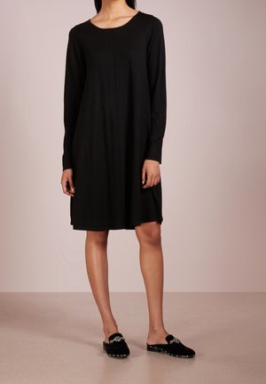 Vrouw die een zwarte jurk met lange mouwen tot op de knie draagt en zwarte slip-on schoenen met zilveren versieringen, staand tegen een effen achtergrond.