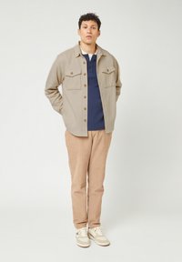 Beige knopenjack met twee borstzakken, een marineblauwe polo eronder, tan kleurige corduroy broek en witte sneakers met grijze accenten.