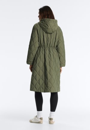 Žena stojící zády, oblečená v dlouhém olivově zeleném prošívaném kabátě s kapucí, černých legínách, bílých ponožkách a béžových teniskách.