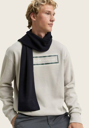 Écharpe en tricot bleu foncé drapée autour du cou, portée avec un sweatshirt gris clair présentant un logo encadré au centre.