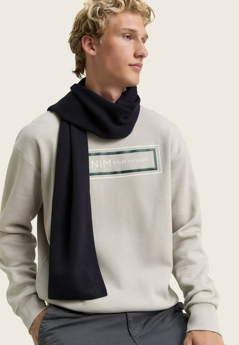 Ciemnoniebieski, dzianinowy szalik owinięty wokół szyi, noszony z jasnoszarym swetrem z centralnie umieszczonym ramkowym logo.