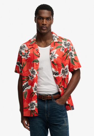 Man wearing open red Hawaiian shirt with white flowers over white tank top, blue jeans, and braided brown belt, standing with one hand in pocket.
