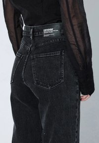 Jeans in denim nero con vita alta, dotati di due tasche posteriori e un'etichetta tessuta. Il tessuto presenta una leggera texture e un aspetto scolorito.