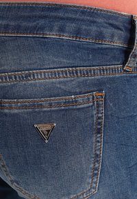 Jeans de mezclilla de un azul oscuro con un bolsillo trasero prominente que presenta un logo triangular metálico. Costuras contrastantes visibles a lo largo de los bordes del bolsillo.
