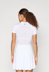 Biała sukienka sportowa z dopasowanym gorsetem, krótkimi rękawami i rozkloszowanym dołem. Posiada perforowane plecy oraz kontrastowy czarny kołnierzyk.