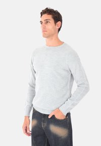 Maglione lavorato a maglia grigio chiaro con collo rotondo e polsini a coste, indossato su jeans di denim scuri con leggera usura.
