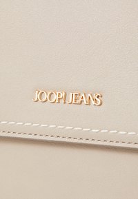 Beige tekstureret materiale med guld "JOOP! JEANS" logo og hvide syninger, der har en glat finish og minimalistisk design.