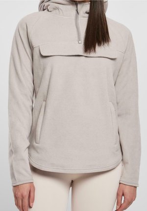 Kvinde iført lys grå fleece-pullover med lynlåslukning foran på kraven og to sidelommer, kombineret med beige bukser.