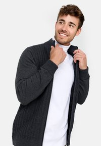 Mørkegrå zip-up cardigan med ribbede teksturer på ærmerne og den øvre del, båret over en hvid turtleneck med høj krave.
