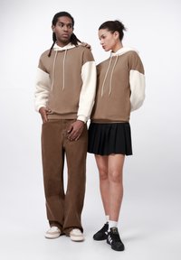 Felpe con cappuccio bicolori marroni con maniche cremose testurizzate, abbinate a pantaloni cargo marroni e una gonna plissettata nera. Calzature: scarpe beige e nere.