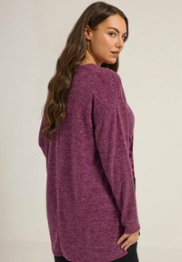 Femme aux longs cheveux bruns portant un pull violet ample à manches longues, regardant par-dessus son épaule contre un fond uni.
