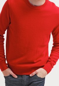 Czerwony sweter z dzianiny z okrągłym dekoltem i długimi rękawami. Żebrowane mankiety i dół, wykonany z miękkiego materiału, zestawiony z ciemnoniebieskimi dżinsami.