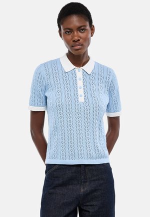 Femme portant un polo en maille bleu clair à manches courtes avec col et poignets blancs, associé à un jean bleu foncé.