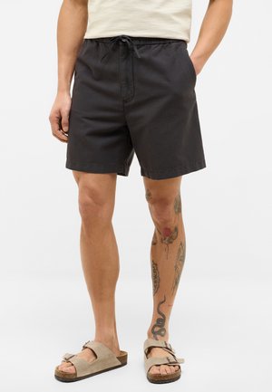 Schwarze Baumwollshorts mit einem elastischen Bund, Vorderentaschen und einer lockeren Passform. Getragen mit beigen Hausschuhsandalen.