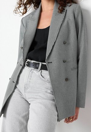 Femme portant un blazer gris à carreaux, un haut noir, un jean gris clair et une ceinture noire, avec des cheveux bouclés partiellement visibles.