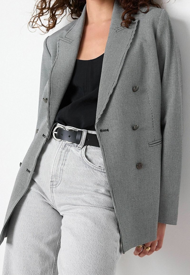 Femme portant un blazer gris à carreaux, un haut noir, un jean gris clair et une ceinture noire, avec des cheveux bouclés partiellement visibles.