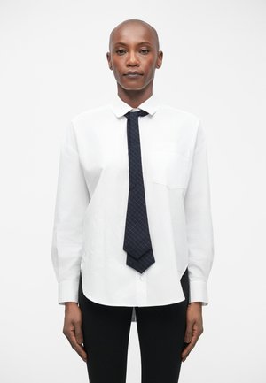 Bijela košulja s dugim rukavima i prednjim džepom, u kombinaciji s tamnoplavom kravatom sa uzorkom mreže. Crne tajice.