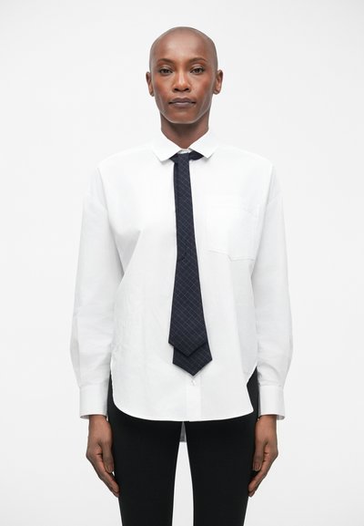 Chemise blanche à manches longues avec une poche frontale, associée à une cravate bleu marine à motif en carreaux. Leggings noirs.
