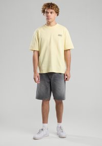 Yellow cotton T-shirt with short sleeves and graphic detail, paired with gray denim shorts and white sneakers. Simple, relaxed fit.