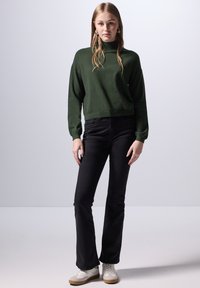Suéter de punto verde oscuro con cuello alto, combinado con jeans negros de campana y zapatillas blancas. Diseño simple con mangas largas.