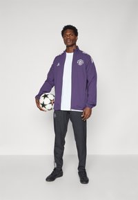 Lila Trainingsjacke mit Logo, weißes Shirt, schwarze Hose mit Logo und schwarze Fußballschuhe, hält einen Fußball. Mit gestreiften Akzenten.