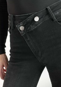 Jean en denim noir à la coupe ajustée, avec deux boutons argentés à la taille et une texture subtile. Poche latérale visible.