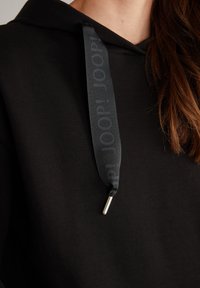 Felpa nera con cappuccio e coulisse, caratterizzata da un nastro nero texturizzato e un puntale in metallo. Il tessuto sembra morbido e presenta una superficie liscia.