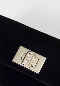 Čierny semišový clutch s lesklým strieborným obdĺžnikovým uzáverom s embosovaným brandingom a kruhovým akcentom. Hladký povrch, minimalistický dizajn.