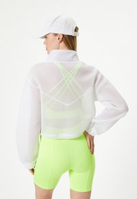 Hvid netcropped jakke med lange ærmer og høj krave, parret med neon grønne cykelshorts. Kappen tilføjer et afslappet sportswear-look.