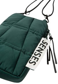 Grünes, gesteppter Beutel mit schwarzem Reißverschluss und Riemen. Verfügt über ein Etikett mit der Aufschrift "SENSES" in fettem Schwarz auf weißem Hintergrund.