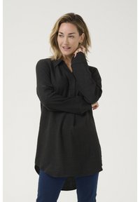 Schwarzes Langarmshirt mit Kragen, strukturiertem Stoff und abgerundetem Saum, kombiniert mit blauen Hosen.