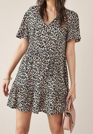 Femme portant une robe courte à imprimé léopard, tenant un petit sac à main rose clair et portant des bracelets dorés au poignet gauche.