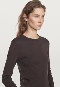 Mørkebrun langærmet skjorte lavet af blødt stof, med rund halsudskæring og tætsiddende design, samt subtile stikninger på kanterne.