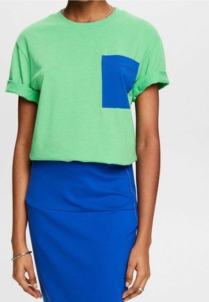Personne portant une chemise à manches courtes vert clair avec une poche bleue et des manches retroussées, ainsi qu'une jupe ajustée bleu vif.