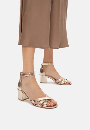 Złote sandały na obcasie typu blok, z krzyżującymi się paskami i paskiem wokół kostki z okrągłą metalową klamrą.