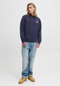 Navy blue cotton hoodie with a kangaroo pocket, adjustable drawstring, and a small red logo label. Paired with light blue jeans and yellow boots.