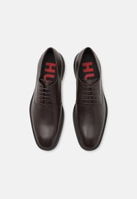 Chaussures de costume en cuir marron avec une finition lisse, bout arrondi et semelle en caoutchouc noir. Comprend des lacets et des semelles intérieures de marque rouges.