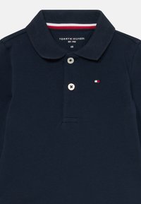 Tamsiai mėlynas polo marškinėliai, pagaminti iš tekstūruoto audinio, su klasikinėmis apykaklėmis, trimis mygtukais ir mažuoju Tommy Hilfiger logotipu raudonai balta spalvomis.