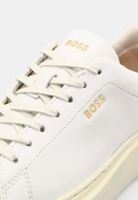 Sneakers in pelle bianca con accenti beige, caratterizzati da una punta rotonda, il logo dorato "BOSS" e lacci piatti. Texture liscia e design minimalista.