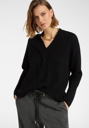 Pull en tricot noir avec un col en V et des manches longues, présentant une coupe ample et une texture côtelée. Associé à un pantalon gris à cordon de serrage.