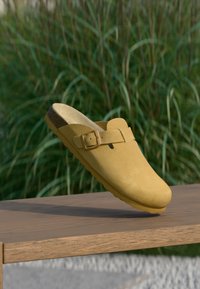 Κίτρινο σουέντ clogs χωρίς κορδόνια με επίπεδη σόλα, ρυθμιζόμενο λουράκι και στρογγυλή μύτη, παρουσιάζεται μπροστά σε φόντο πράσινου γρασιδιού.