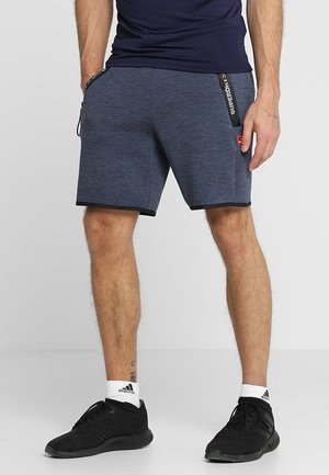 Man draagt marineblauwe sportshorts met zwarte biezen, een marineblauw shirt, witte Adidas-sokken en zwarte sneakers, staand met handen in de zakken.