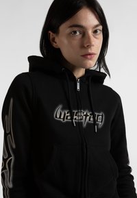 Črna zip-up kapuca iz mehkega blaga, z sprednjim žepom in grafičnim besedilom "wasted" v srebrni barvi. Oblikovanje vključuje abstraktne akcentne detajle na rokavih.