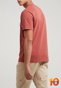 Uomo che indossa una maglietta a maniche corte rossa sbiadita e pantaloni beige ricamati con piccole figure rosse, in piedi davanti a uno sfondo semplice.