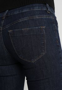 Los jeans de mezclilla oscura presentan un diseño ajustado con una textura suave, costuras sutiles y una forma clásica de bolsillo trasero.