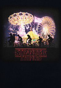 Juodas marškinėlis su siluetu figūrų, važiuojančių dviračiais, ryškūs karnavaliniai atrakcionai fone, su raudonu užrašu "STRANGER THINGS".