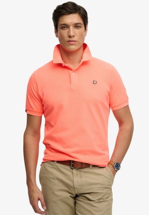 Mand iført koralfarvet kortærmet polo med opstående krave, beige bukser, brunt bælte og armbåndsur, stående med den ene hånd i lommen.