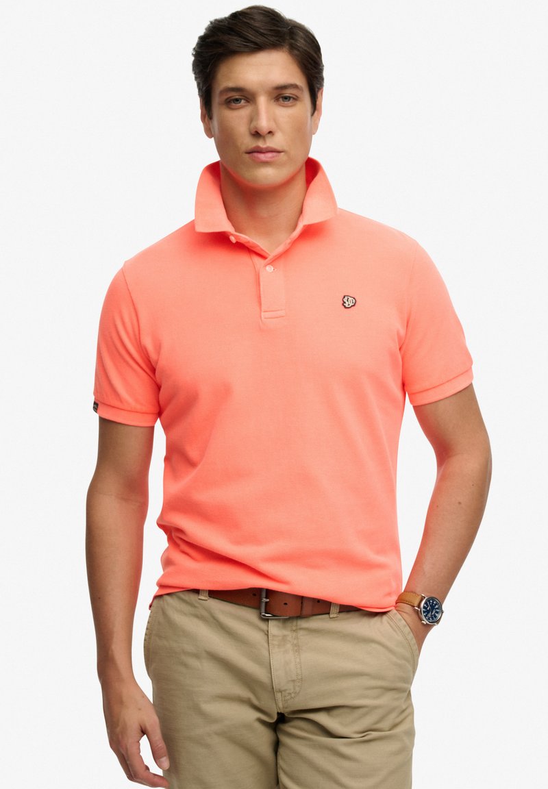 Homme portant un polo corail à manches courtes avec col relevé, pantalon beige, ceinture marron et montre-bracelet, debout avec une main dans la poche.