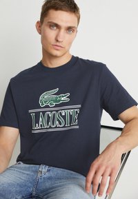 Camiseta de algodón azul marino con un logo de cocodrilo verde y el texto "LACOSTE" debajo. Mangas cortas y cuello redondo. Ajuste casual.