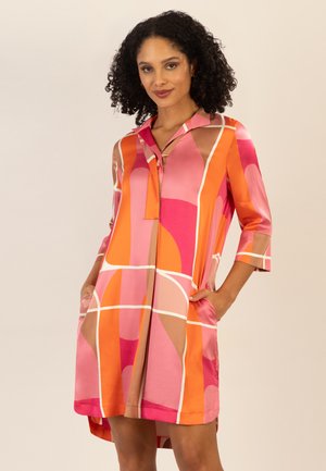 Frau trägt ein lebhaftes Kleid mit geometrischem Muster in Pink, Orange und Beige, mit dreiviertel Ärmeln und Taschen, steht vor einem beigen Hintergrund.